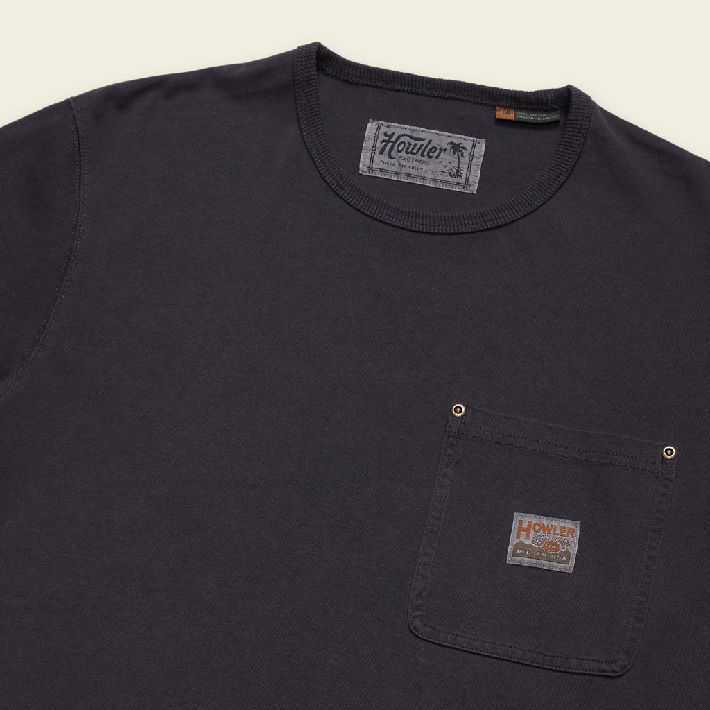 A close-up of a dark charcoal gray crewneck pocket t-shirt by Howler, featuring a chest pocket with brass snap buttons and a branded fabric label.