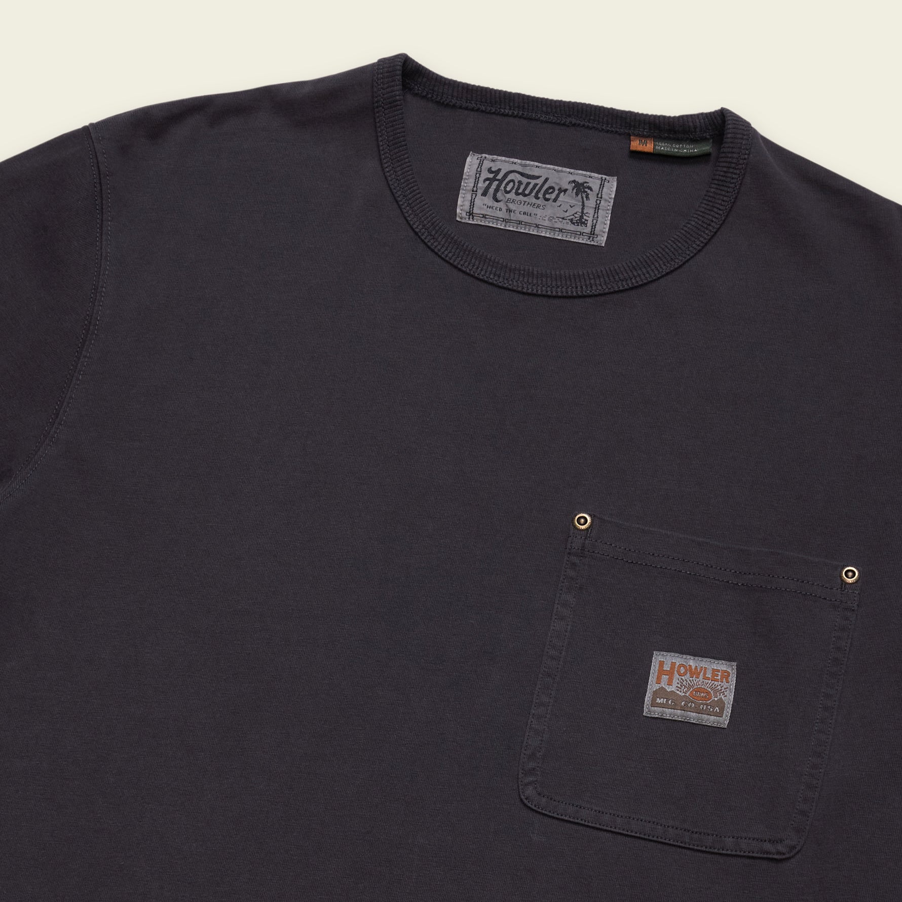 A close-up of a dark charcoal gray crewneck pocket t-shirt by Howler, featuring a chest pocket with brass snap buttons and a branded fabric label.