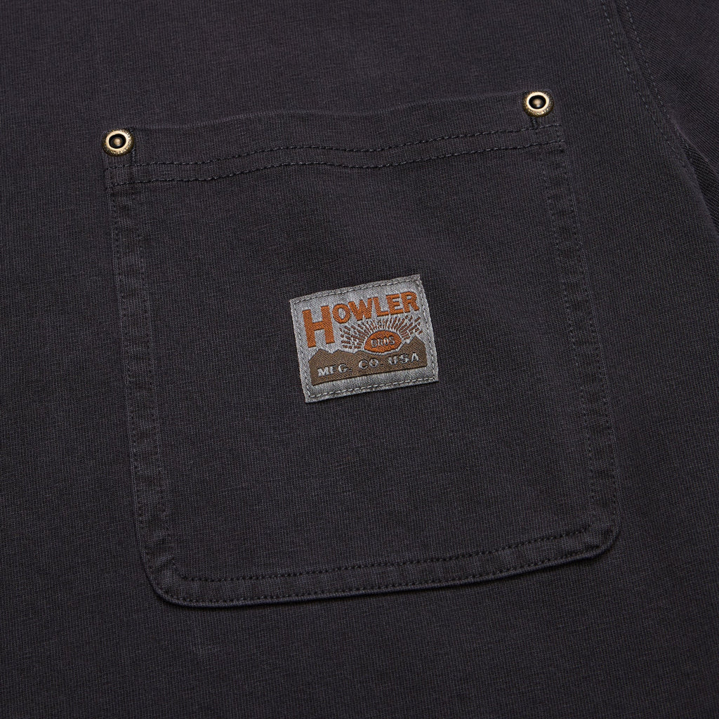 Close-up of a dark charcoal gray or black canvas work shirt pocket with brass snap buttons and a vintage-style 'HOWLER' brand patch.