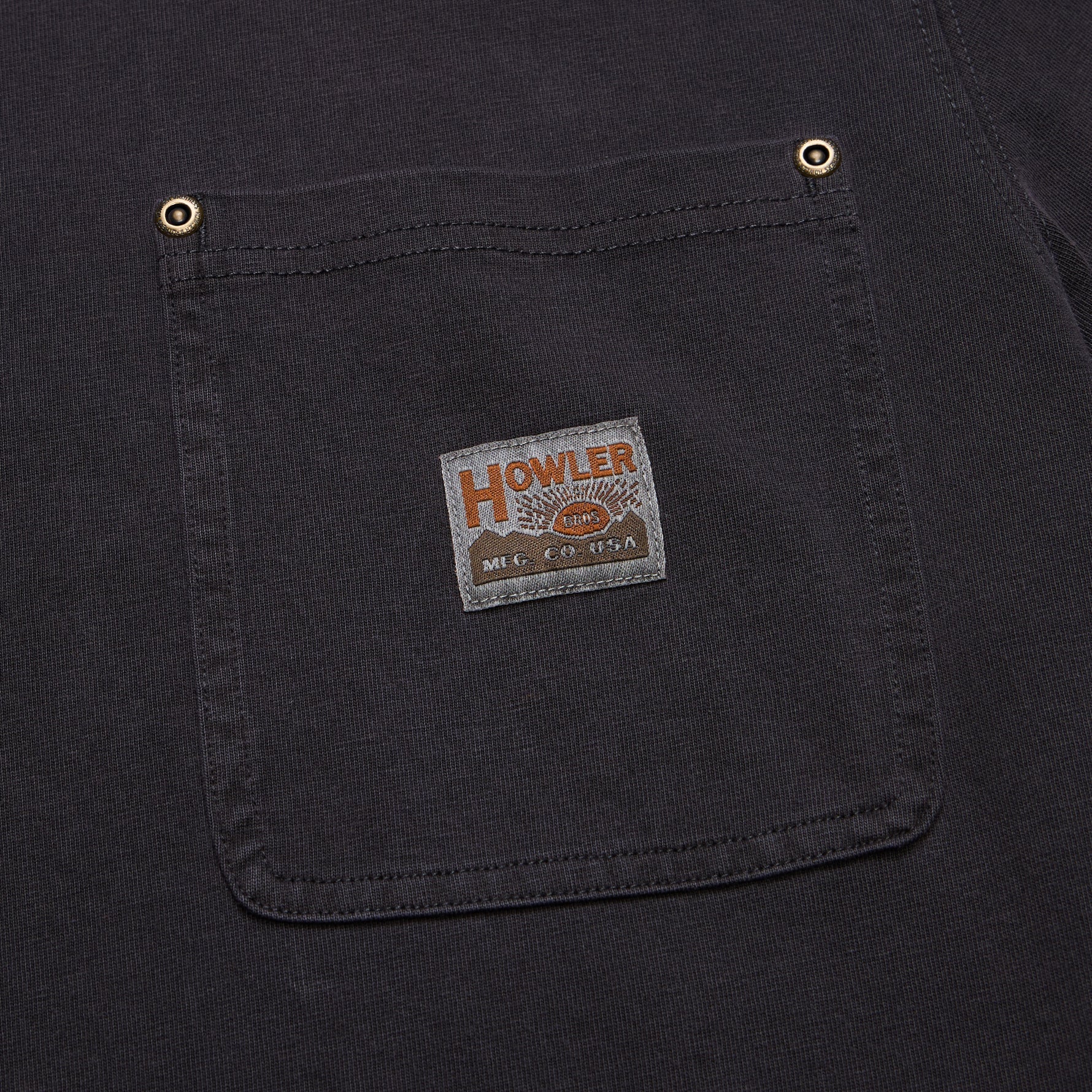 Close-up of a dark charcoal gray or black canvas work shirt pocket with brass snap buttons and a vintage-style 'HOWLER' brand patch.