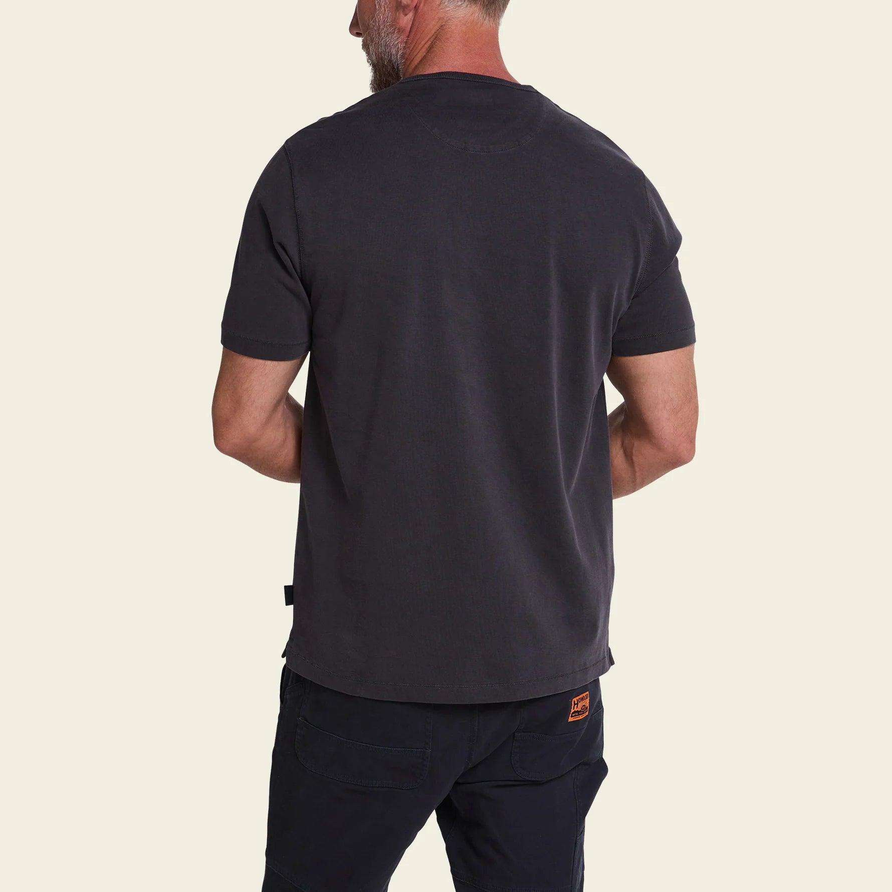 A man shown from the back wearing a dark charcoal gray short-sleeve t-shirt and dark pants, against a solid light beige background.