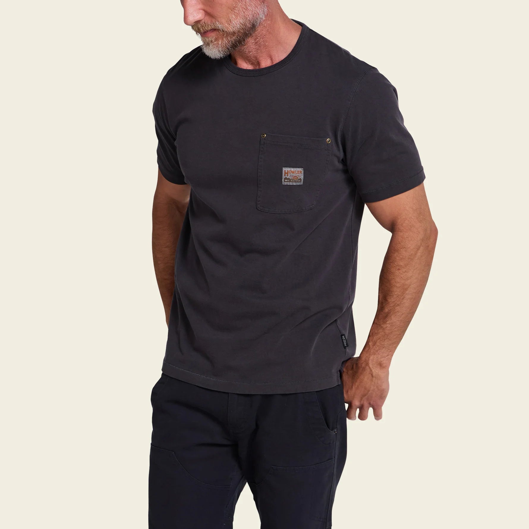 A man wearing a dark charcoal gray short-sleeve pocket t-shirt and dark pants, shown from the waist up against a plain white background.