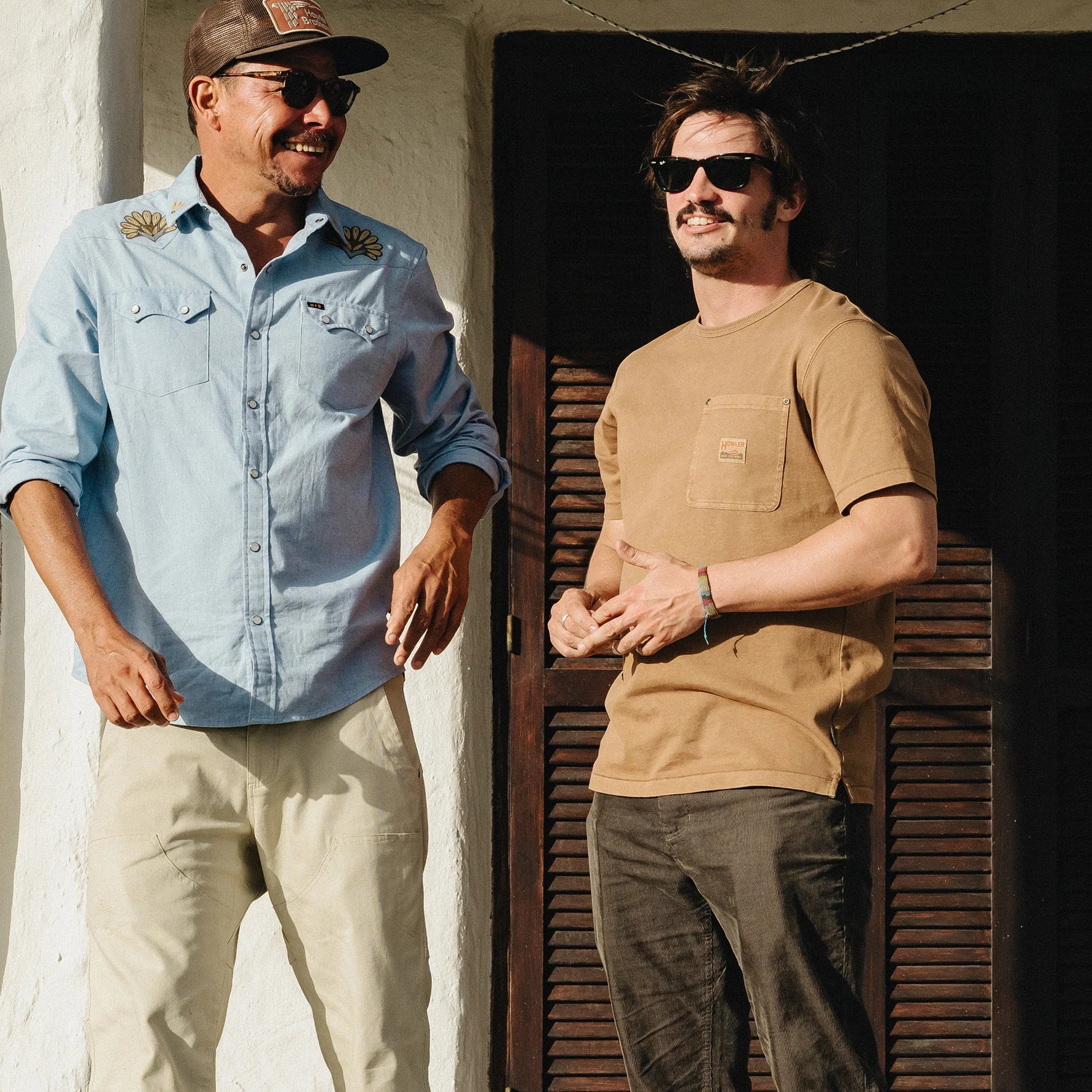 Two men standing outdoors in front of a white stucco wall and dark wooden door. The man on the left wears a light blue button-up shirt, beige pants, sunglasses, and a cap.