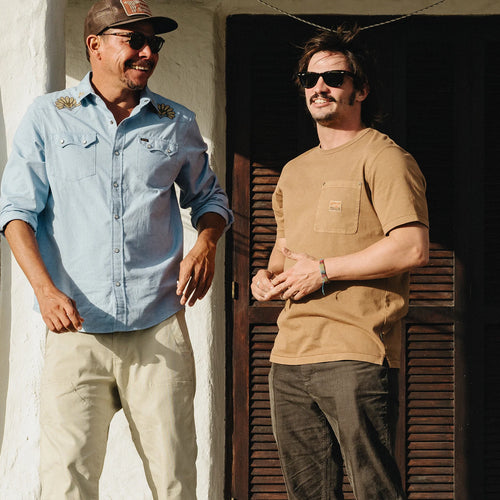 Two men standing outdoors in front of a white stucco wall and dark wooden door. The man on the left wears a light blue button-up shirt, beige pants, sunglasses, and a cap.