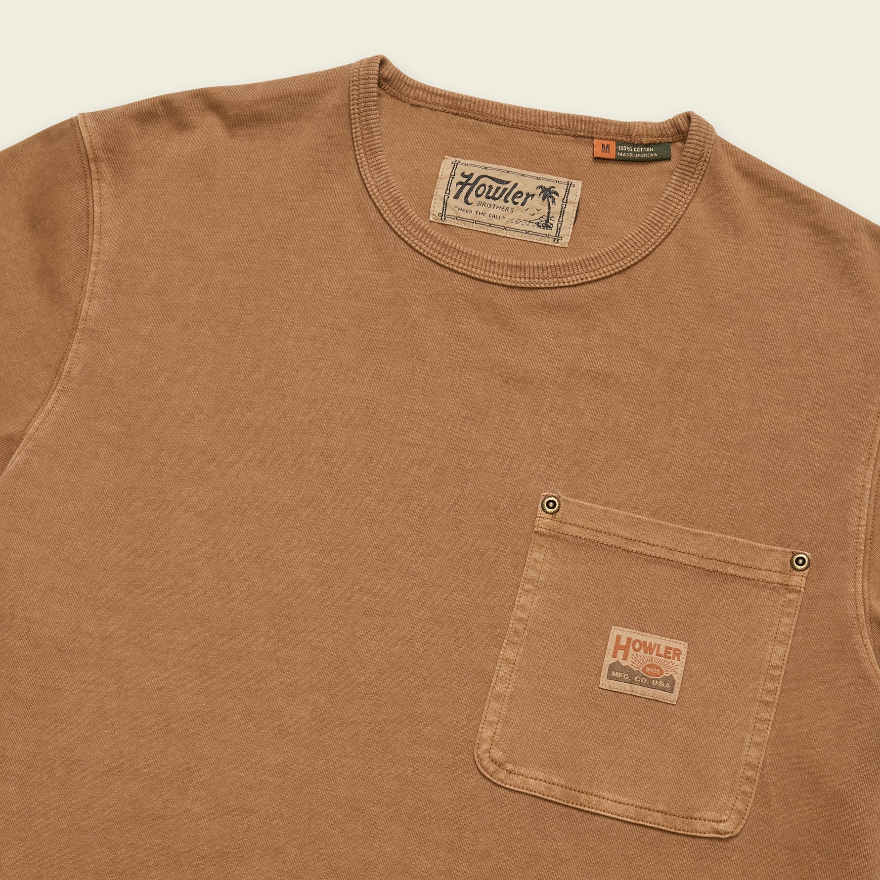 A close-up of a brown cotton crewneck t-shirt with a chest pocket, featuring a visible Howler brand label on the pocket and inner neck tag.