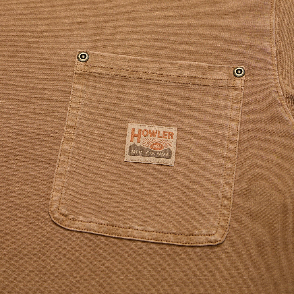 Close-up of a tan work shirt pocket with metal snap buttons and a central sewn-on brand patch reading Howler.