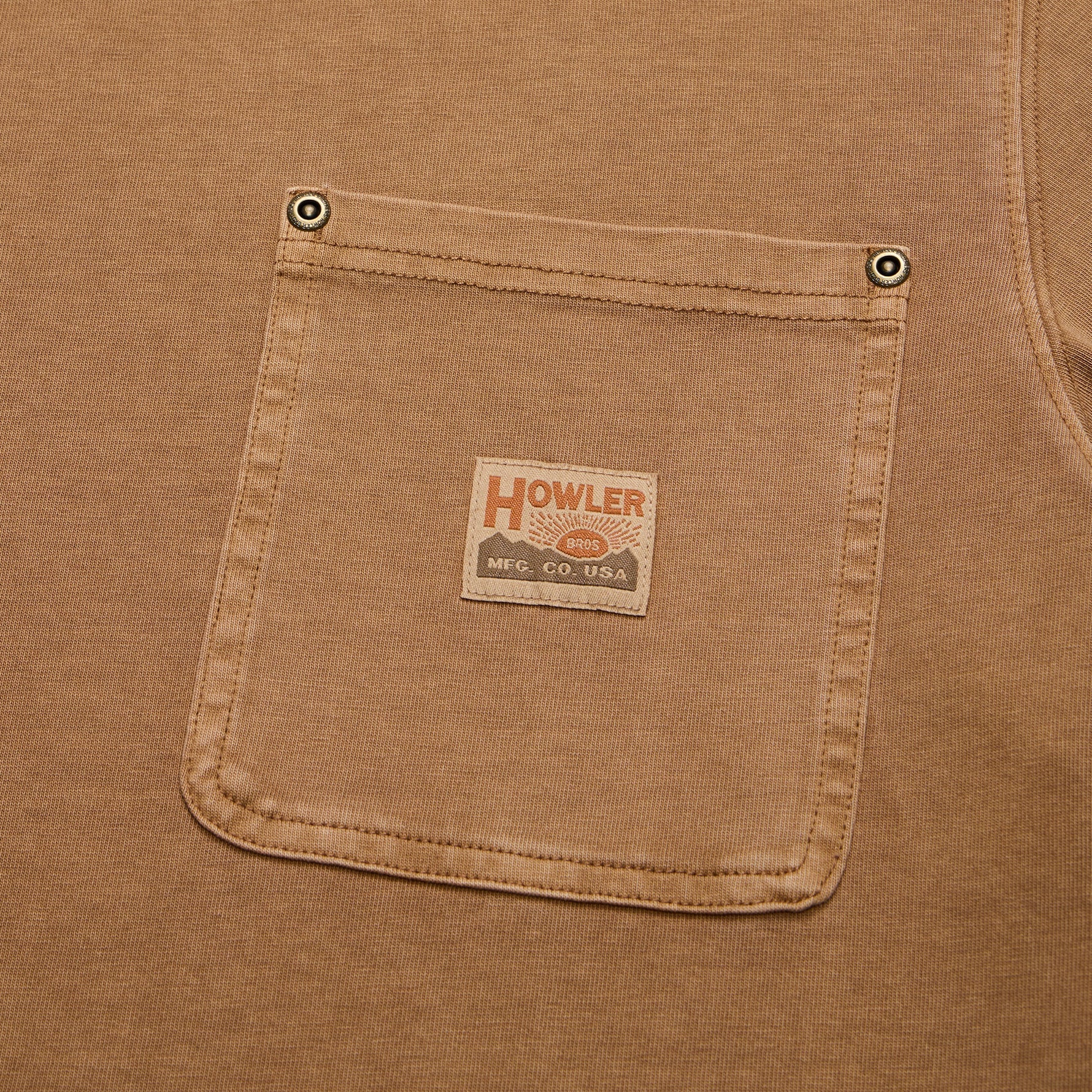Close-up of a tan work shirt pocket with metal snap buttons and a central sewn-on brand patch reading Howler.