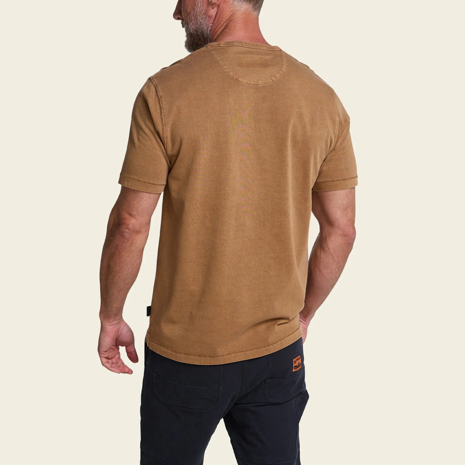 A man shown from the back wearing a tan crewneck t-shirt and dark navy pants against a light cream background.