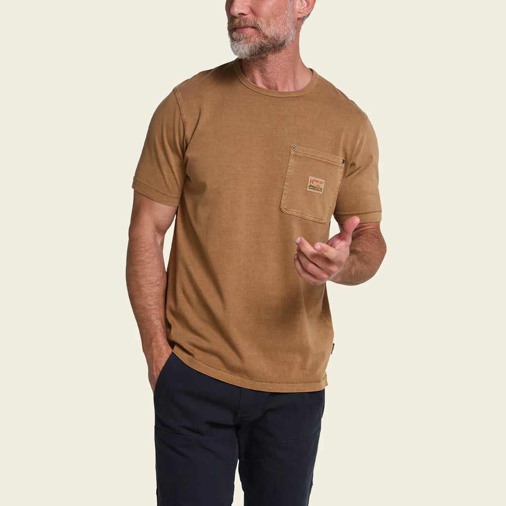 A man wearing a tan short-sleeve crewneck t-shirt with a chest pocket, paired with dark pants, shown against a plain white background.