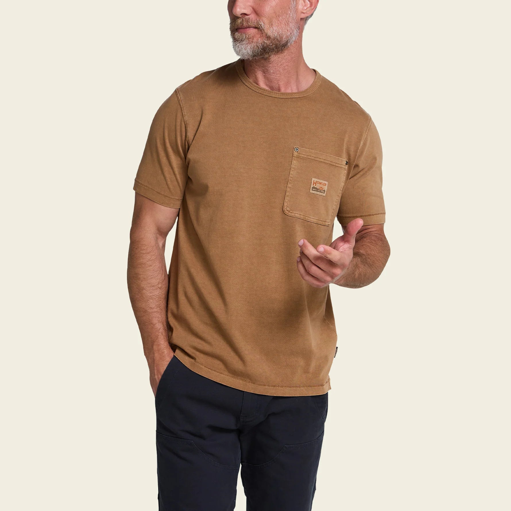 A man wearing a tan short-sleeve crewneck t-shirt with a chest pocket, paired with dark pants, shown against a plain white background.
