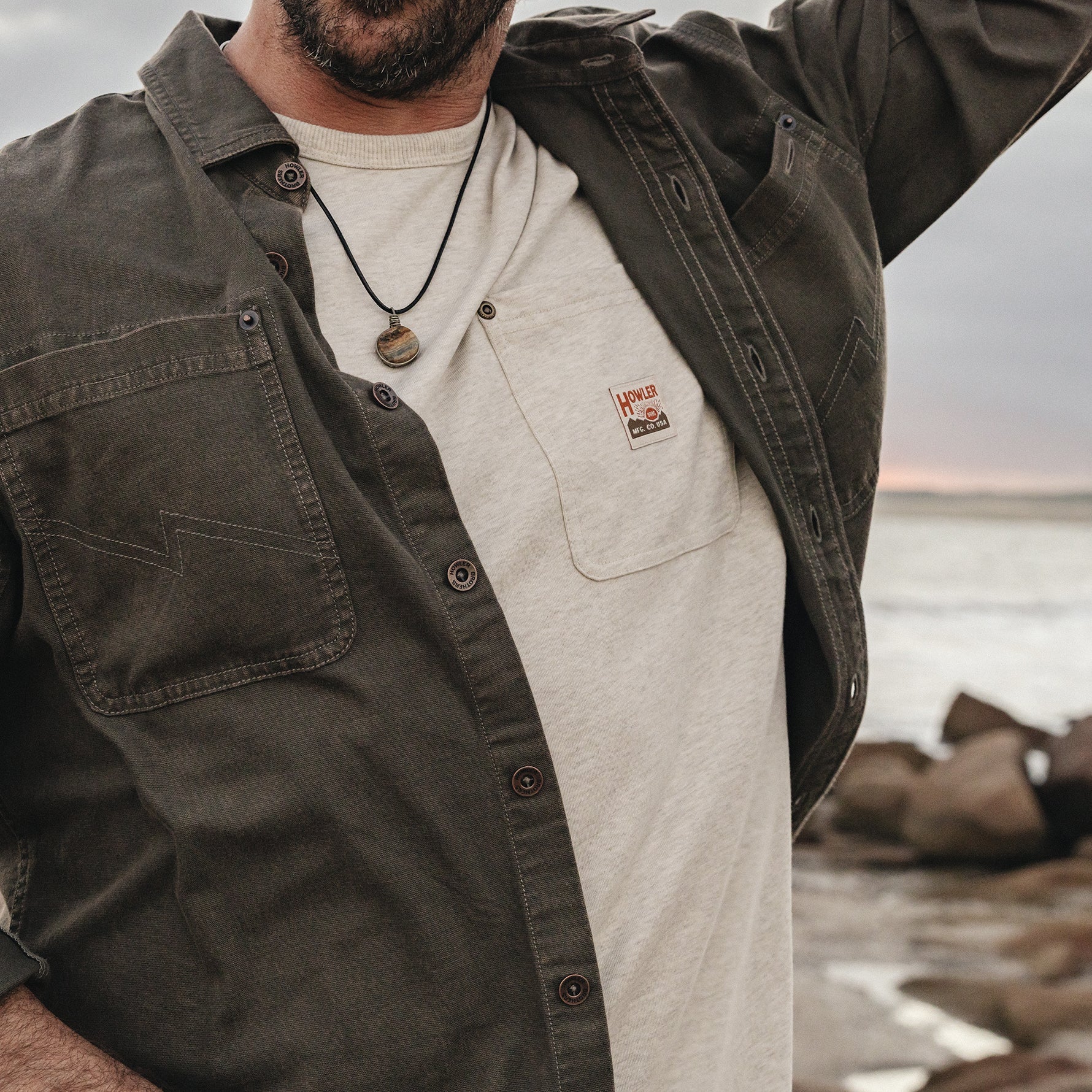 A man wearing an olive green button-up overshirt over a light beige t-shirt with a small red and white 'Hemp' brand patch on the chest pocket, set against a blurred outdoor coastal background.