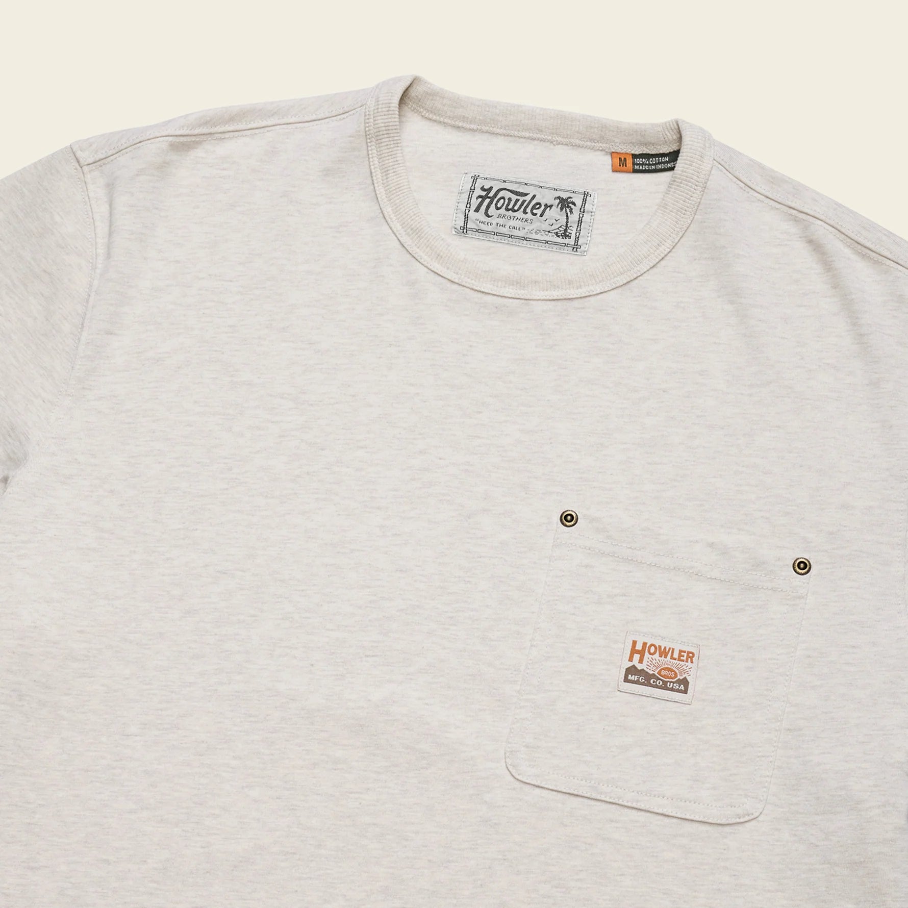 A close-up of a light heather gray crewneck t-shirt with a chest pocket featuring two metal snap buttons and a small Howler brand patch.