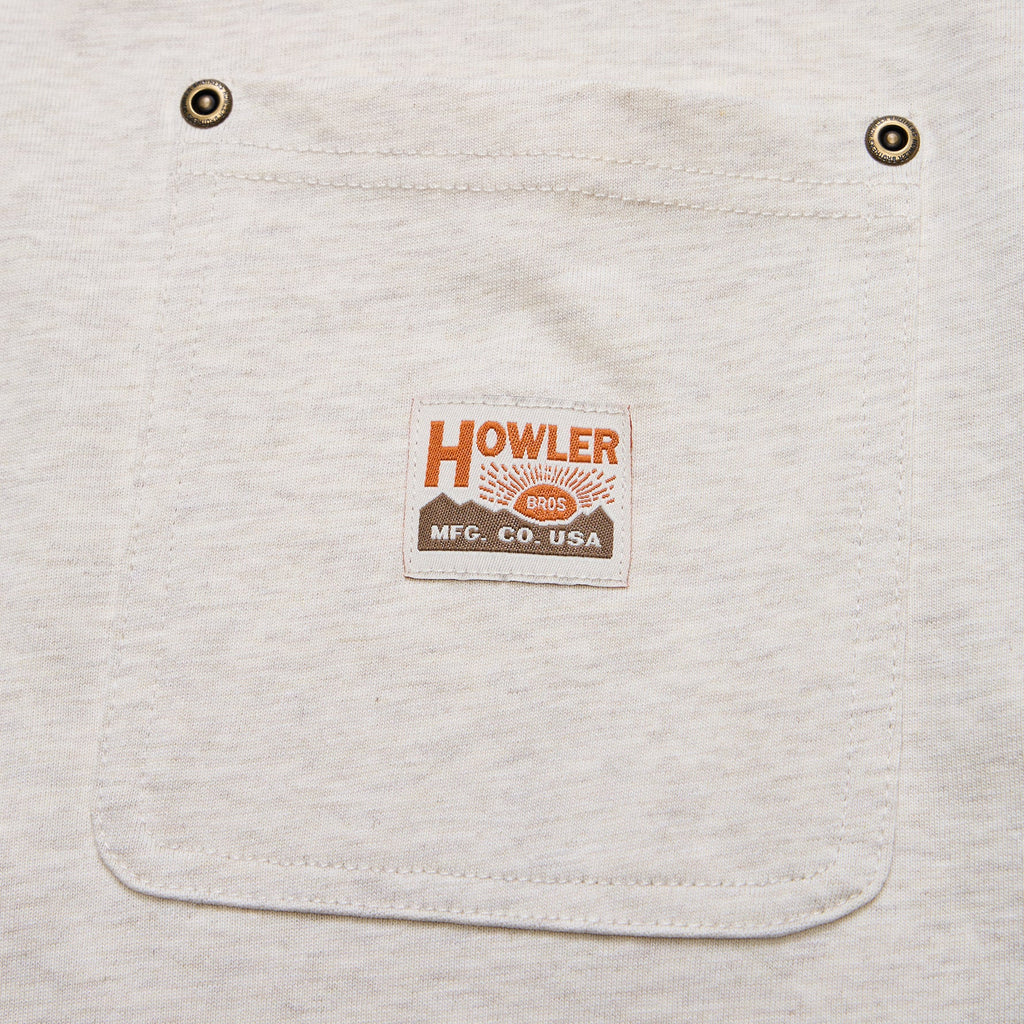 Close-up of a light heather gray fabric pocket with two metal grommets and a centered rectangular brand patch reading HOWLER MFG. CO.