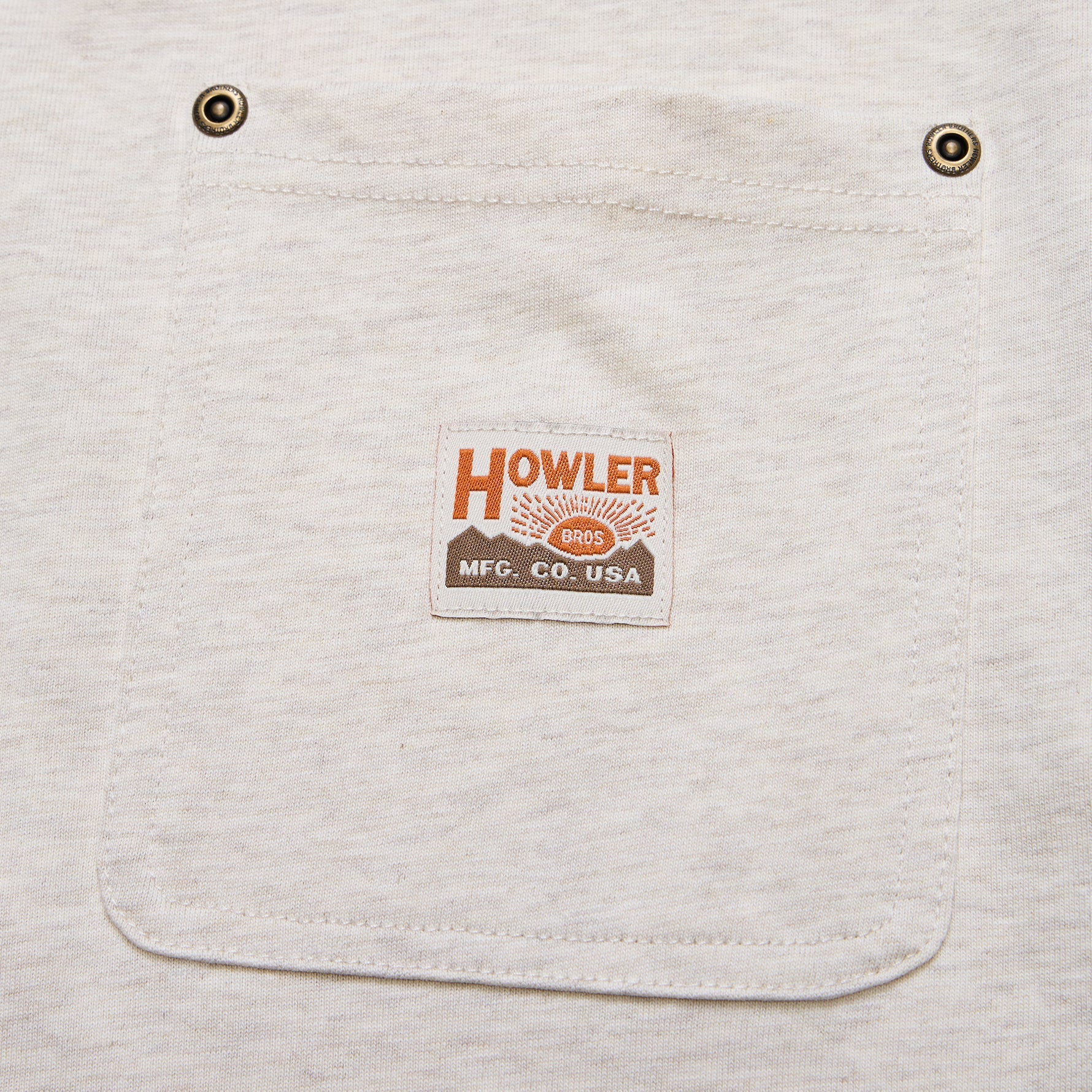 Close-up of a light heather gray fabric pocket with two metal grommets and a centered rectangular brand patch reading HOWLER MFG. CO.