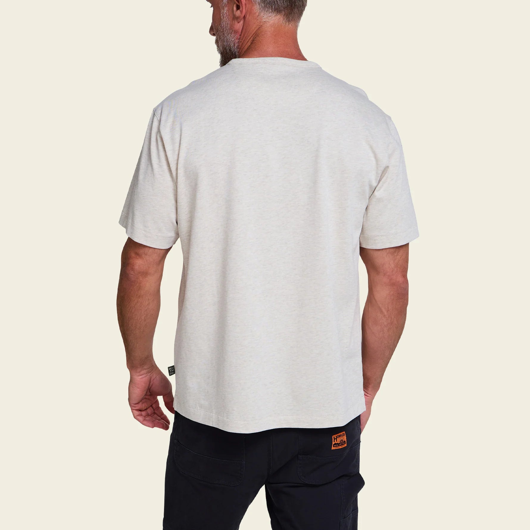 A man viewed from the back wearing a light heather gray crewneck t-shirt and dark pants, set against a solid cream background.