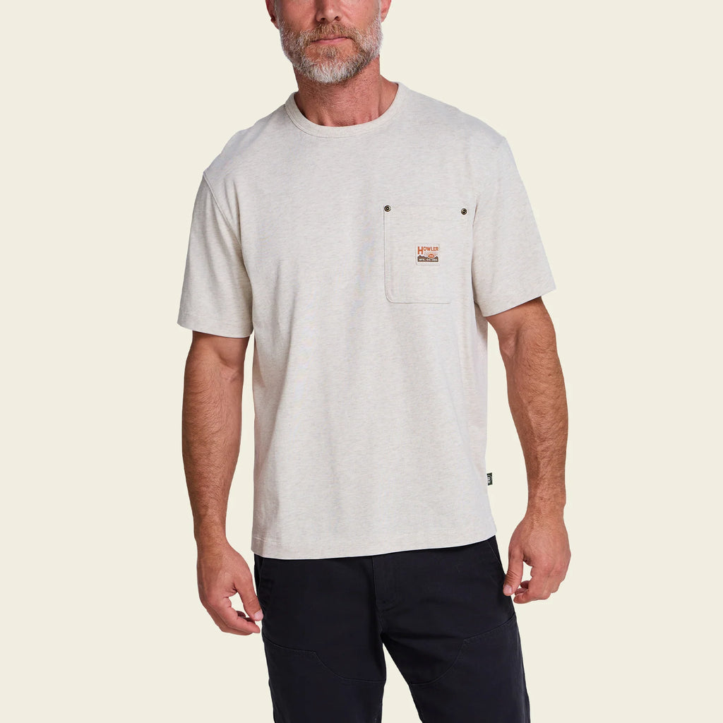 A man wearing a light beige short-sleeve crewneck t-shirt with a chest pocket and a small orange and white logo patch, paired with dark pants, against a solid light beige background.