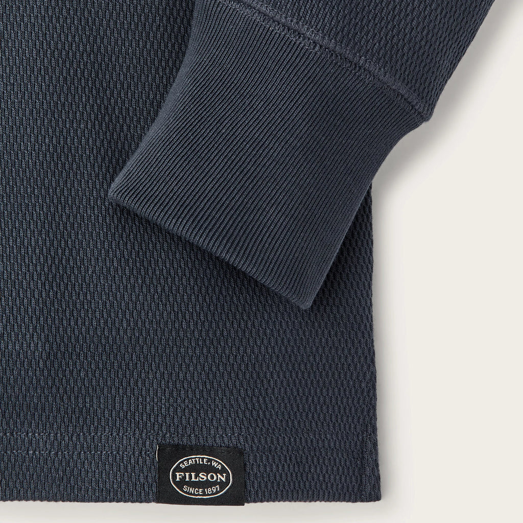 Close-up of a dark navy textured knit garment with a folded cuff and a small black Pilson label near the hem, shown on a light neutral background.