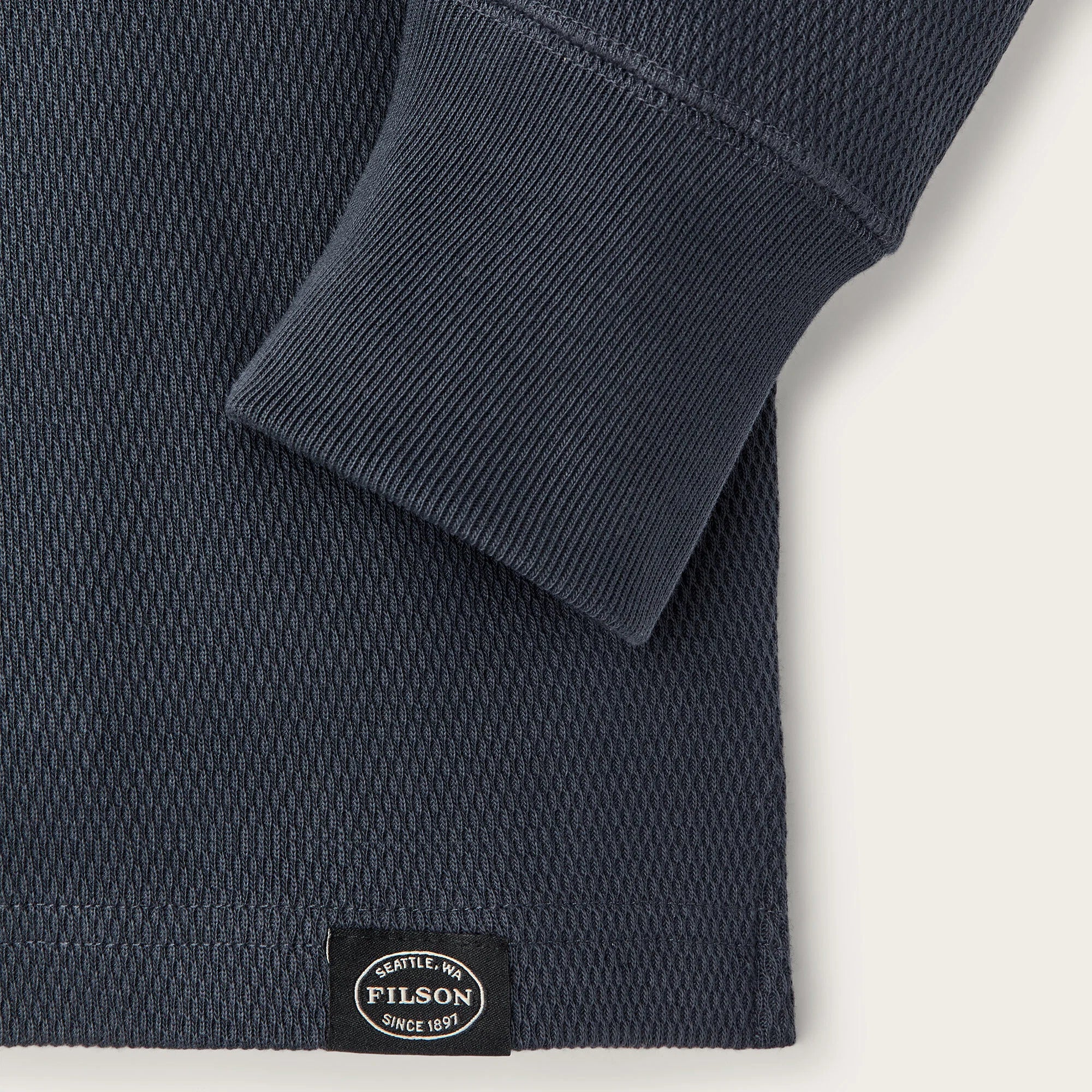 Close-up of a dark navy textured knit garment with a folded cuff and a small black Pilson label near the hem, shown on a light neutral background.