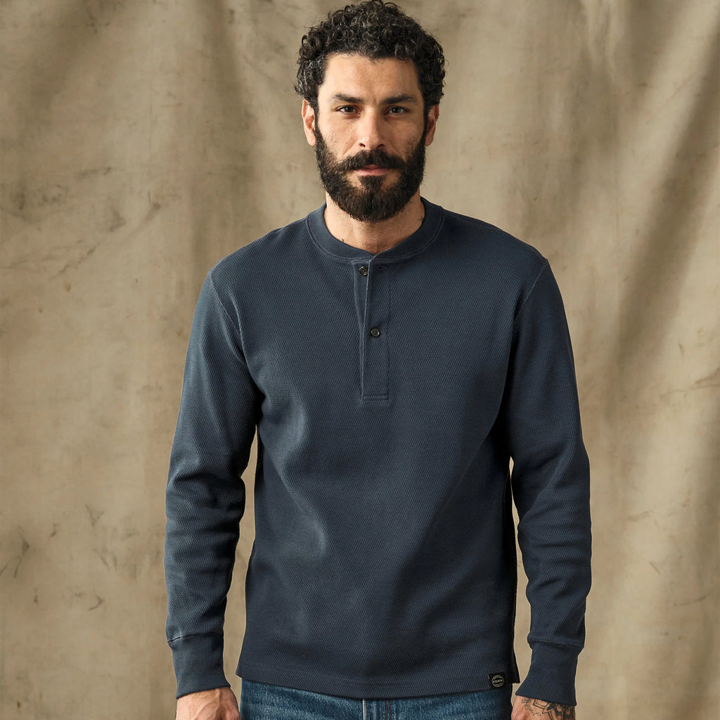 A man with a beard wearing a dark navy blue long-sleeve henley shirt, standing against a textured beige backdrop.