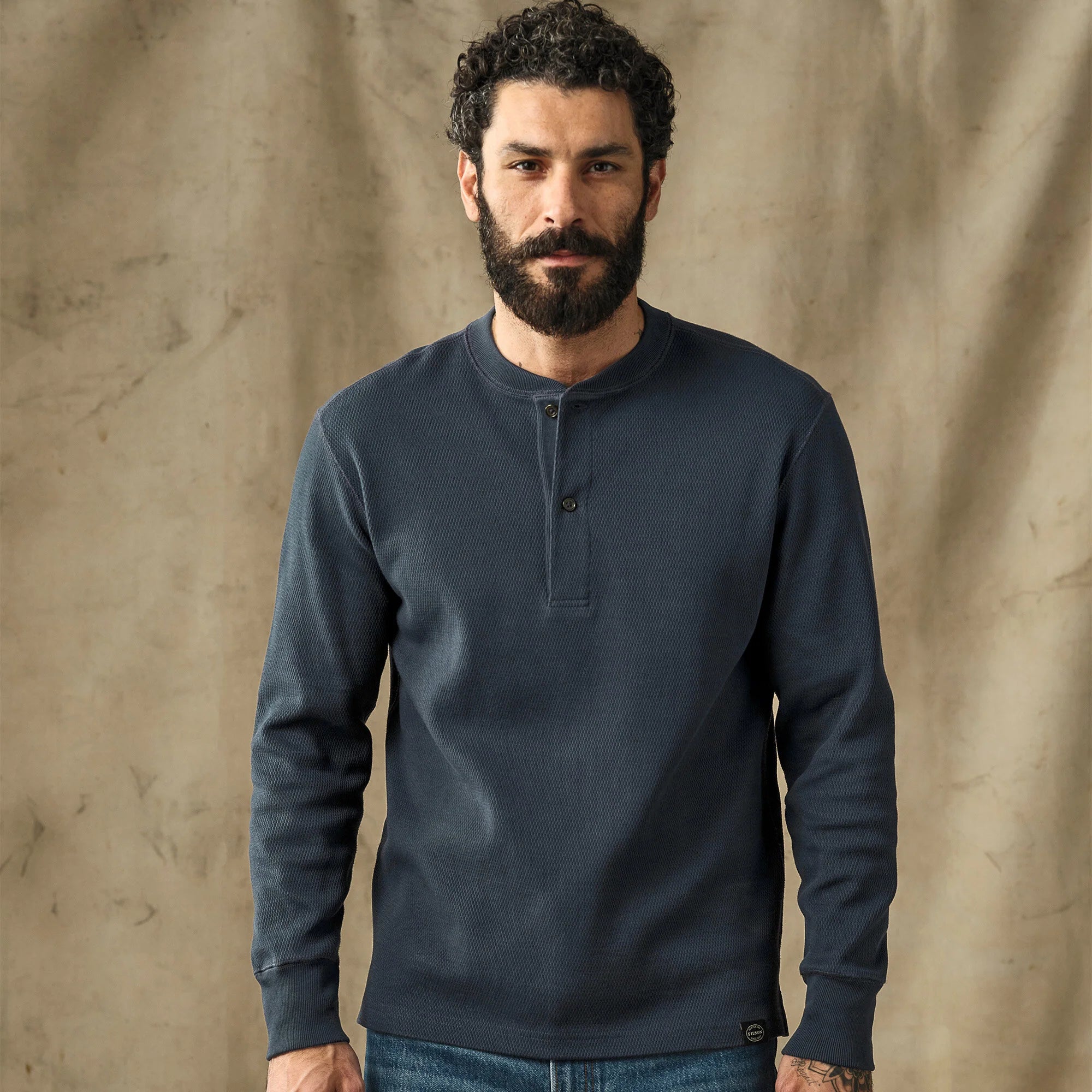 A man with a beard wearing a dark navy blue long-sleeve henley shirt, standing against a textured beige backdrop.