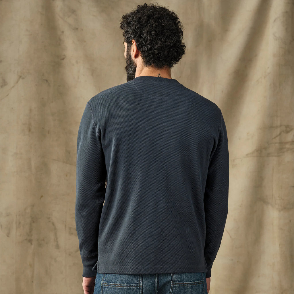 A man with curly dark hair and a beard is shown from behind wearing a dark charcoal gray long-sleeve crewneck shirt and blue jeans, standing against a textured beige backdrop.