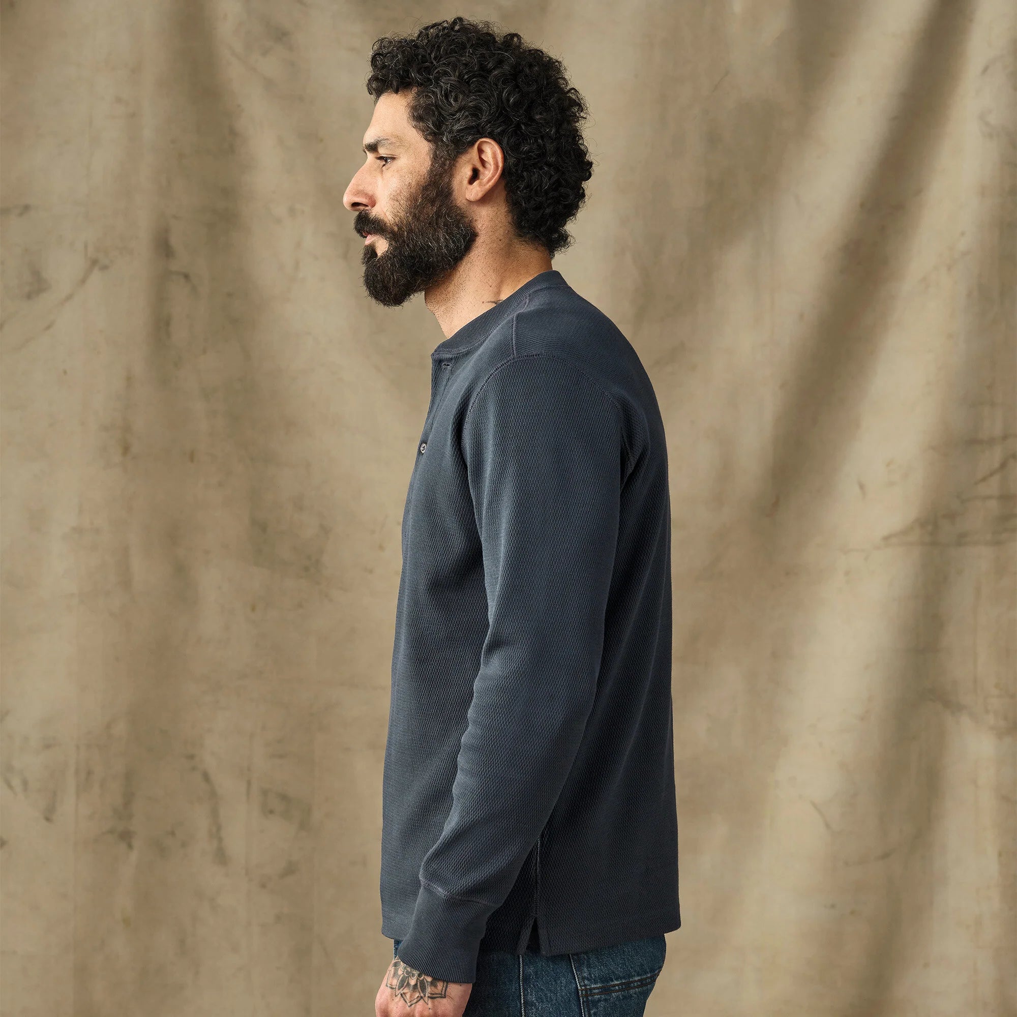 A man with curly black hair and a beard is shown in profile wearing a dark slate blue long-sleeve henley shirt and blue jeans, standing against a textured beige fabric backdrop.