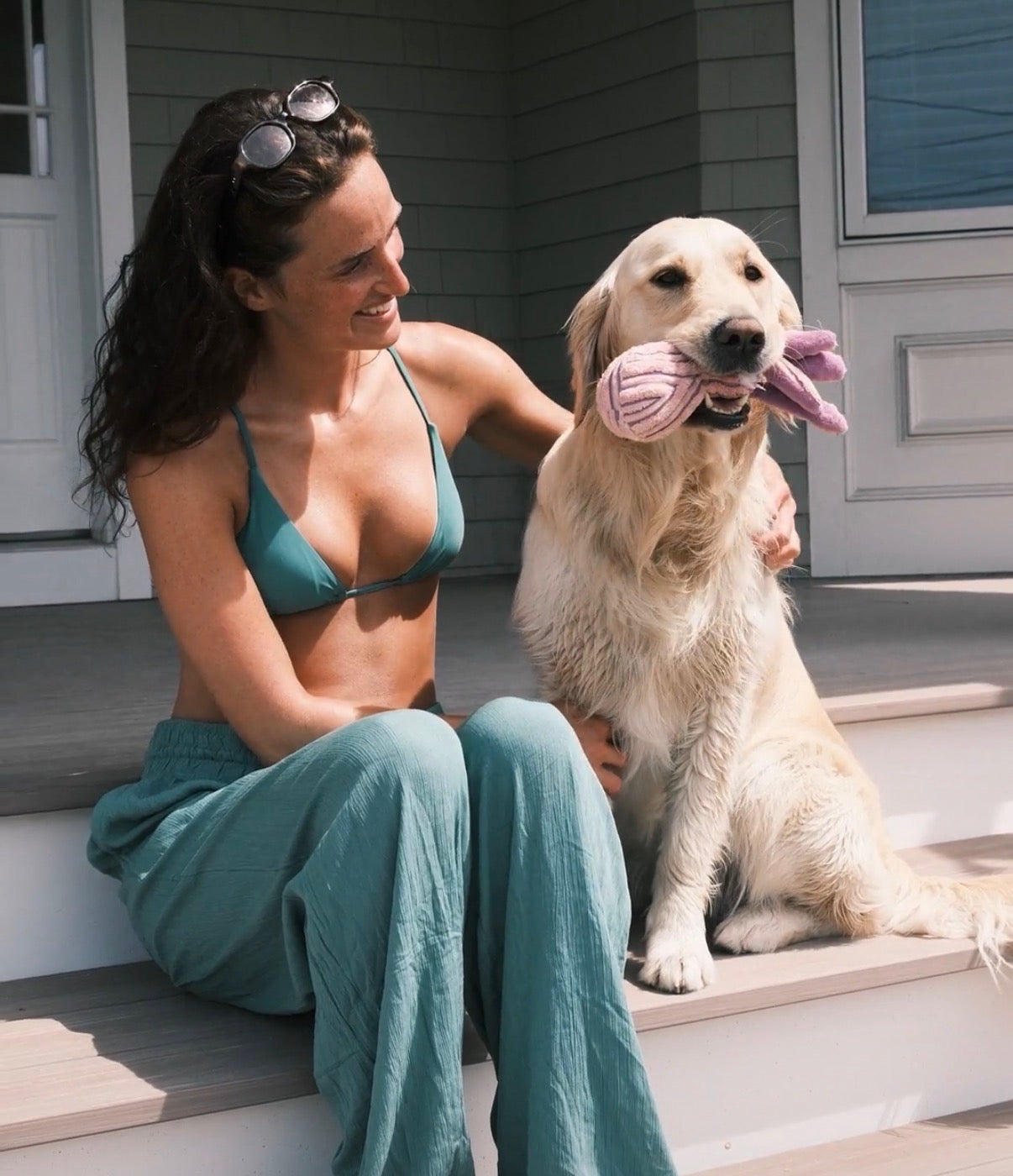 Woman in green bikini and pants sitting on steps with a golden retriever holding a pink toy.