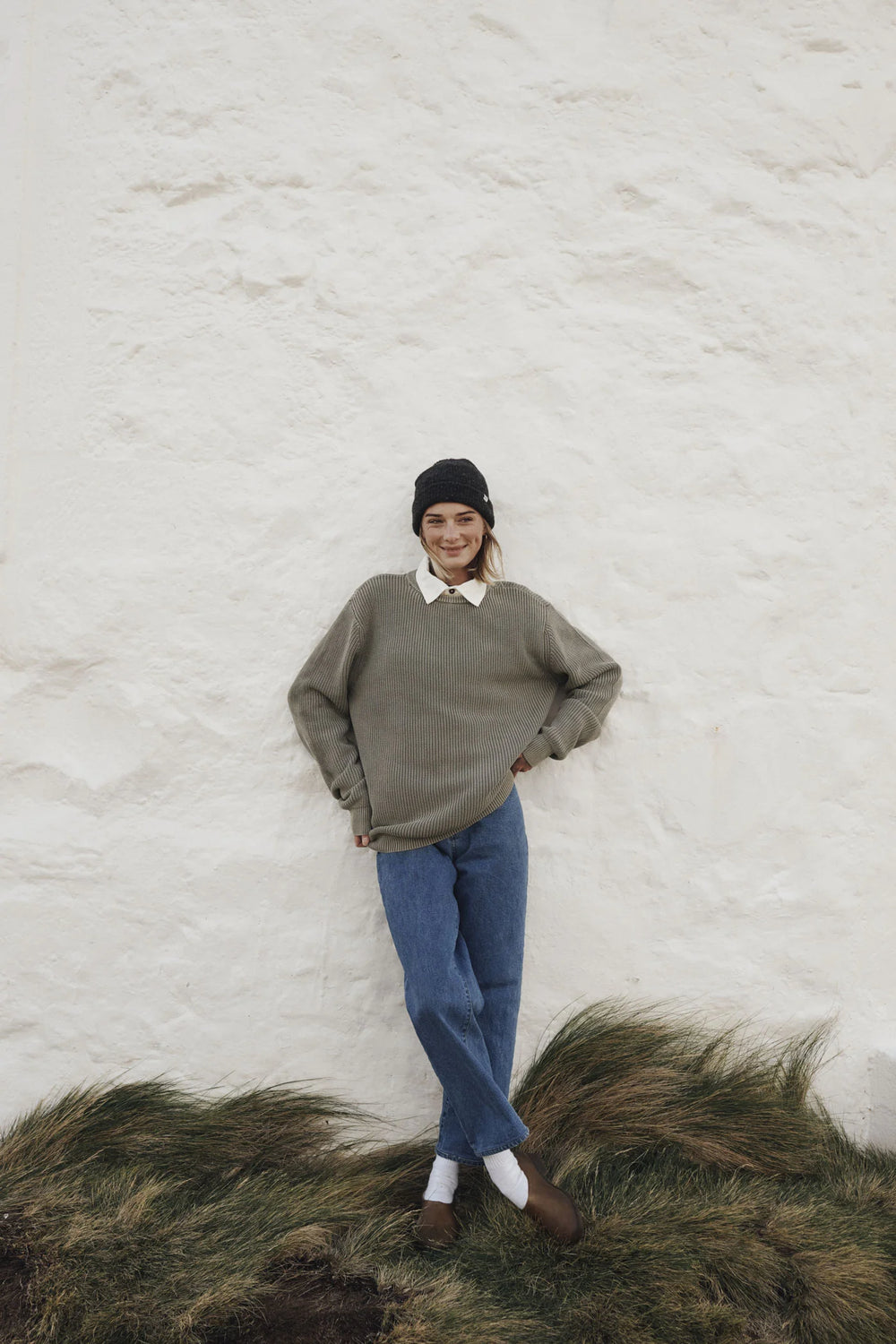 Person wearing a green sweater and blue jeans standing against a white textured wall with grass at the bottom.