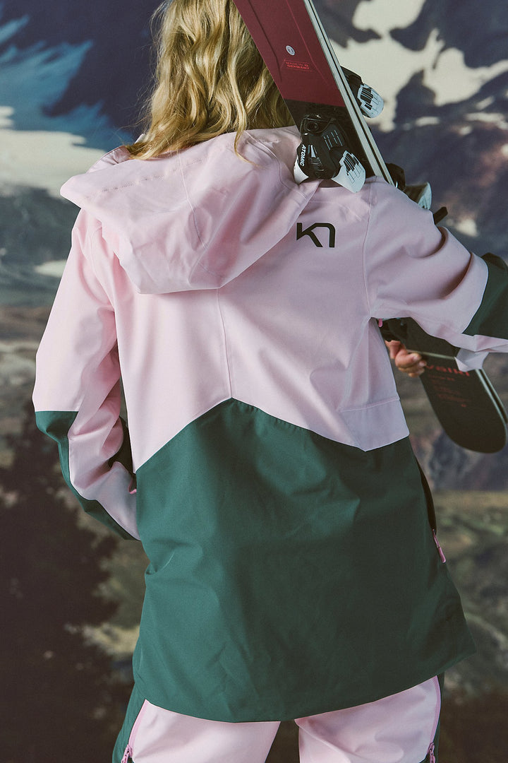 Person wearing a pink and green jacket holding skis with a mountainous background