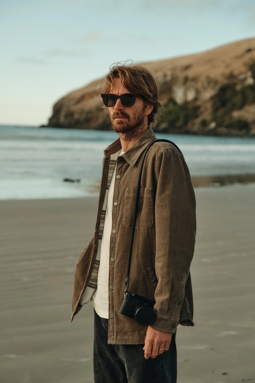 Man standing on a beach wearing sunglasses and a jacket with a scenic background