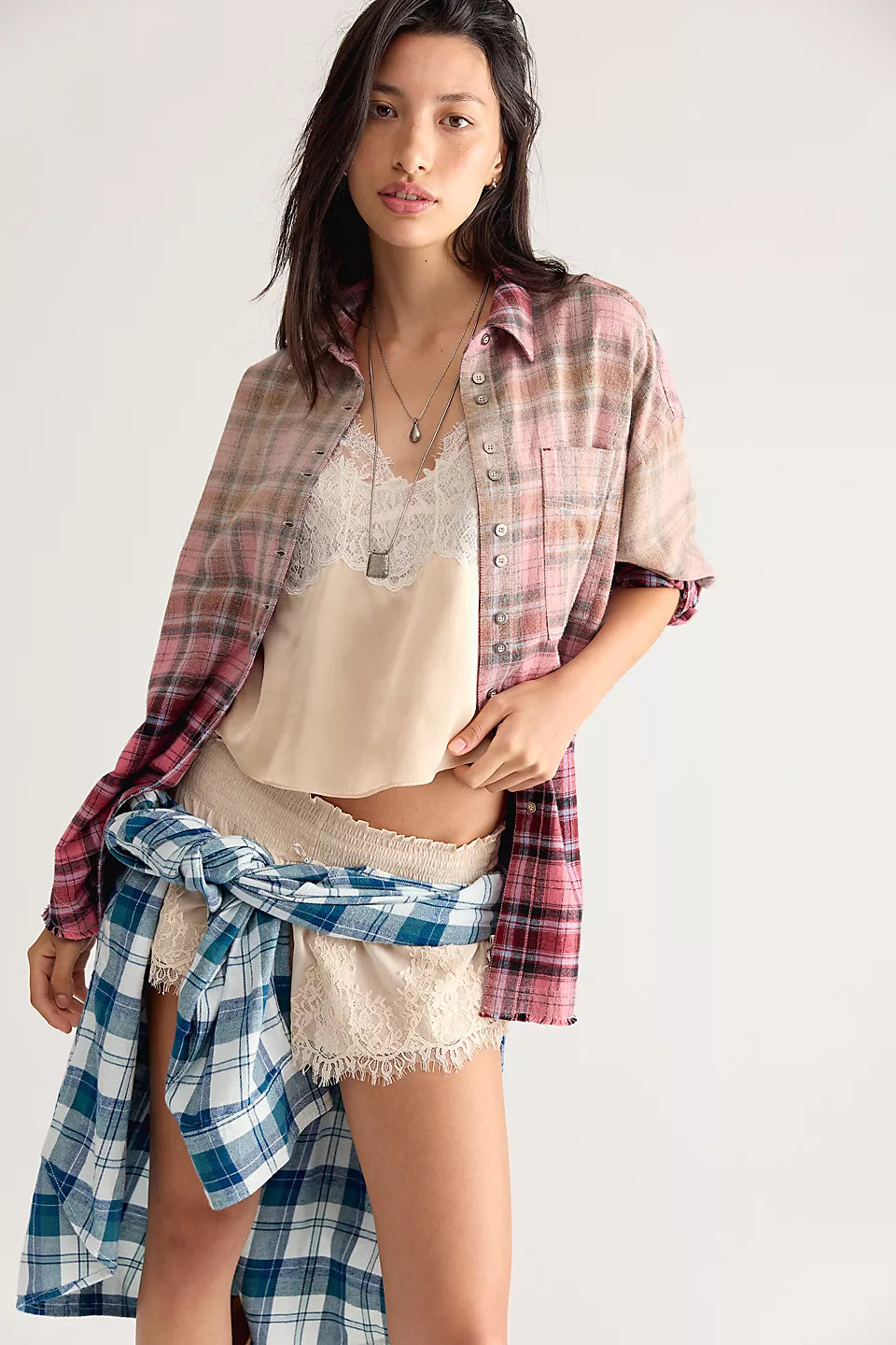 Woman modeling pink and beige plaid shirt, open at the front and sleeves rolled up paired with cream lacy tank top and shorts with blue and white plaid shirt tied around her waist. 