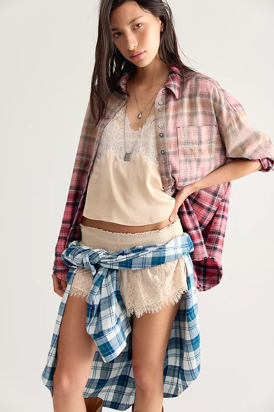 Woman modeling pink and beige plaid shirt, open at the front and sleeves rolled up paired with cream lacy tank top and shorts with blue and white plaid shirt tied around her waist. 