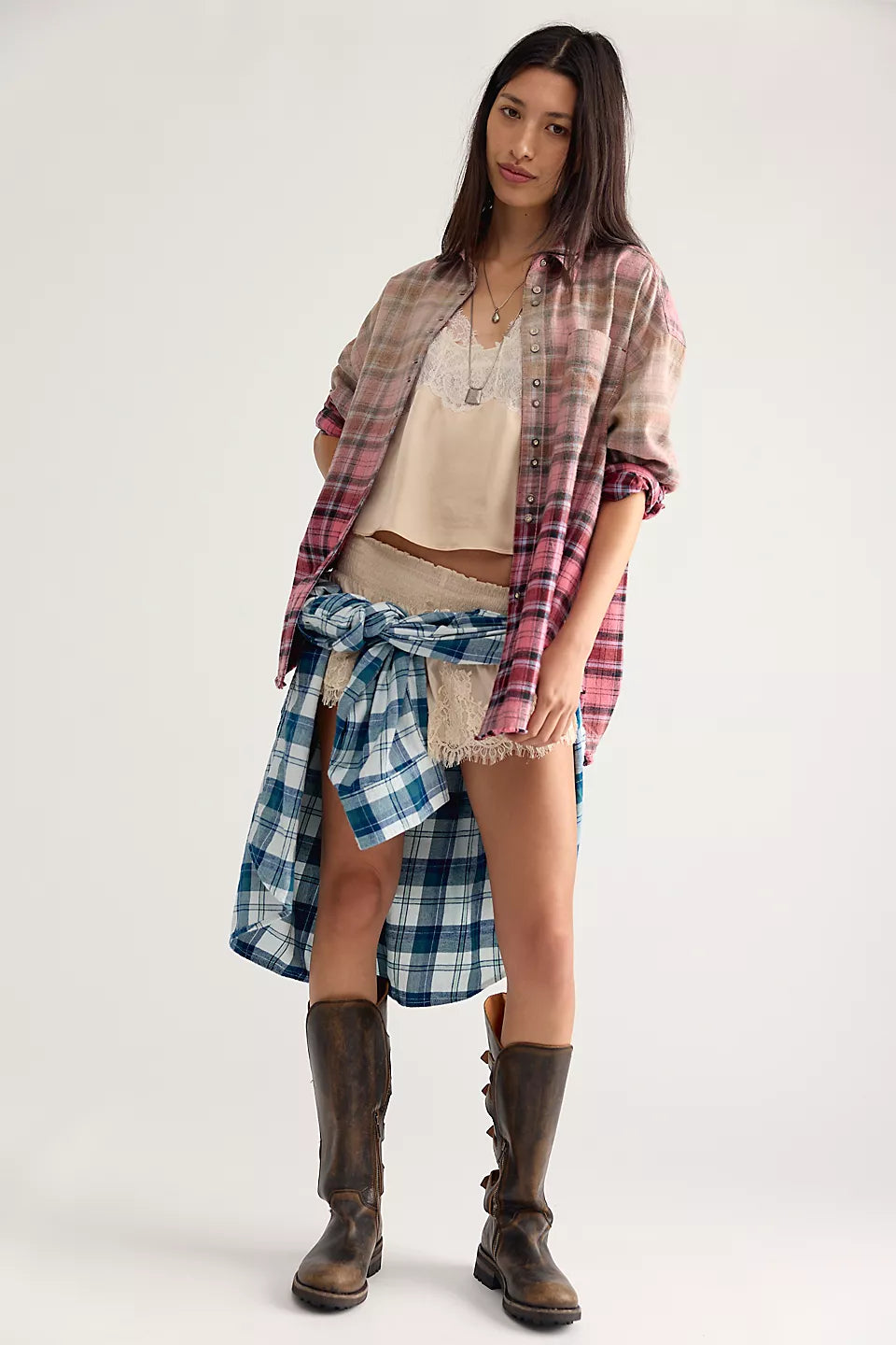 Woman modeling pink and beige plaid shirt, open at the front and sleeves rolled up paired with cream lacy tank top and shorts with blue and white plaid shirt tied around her waist and brown knee high boots.