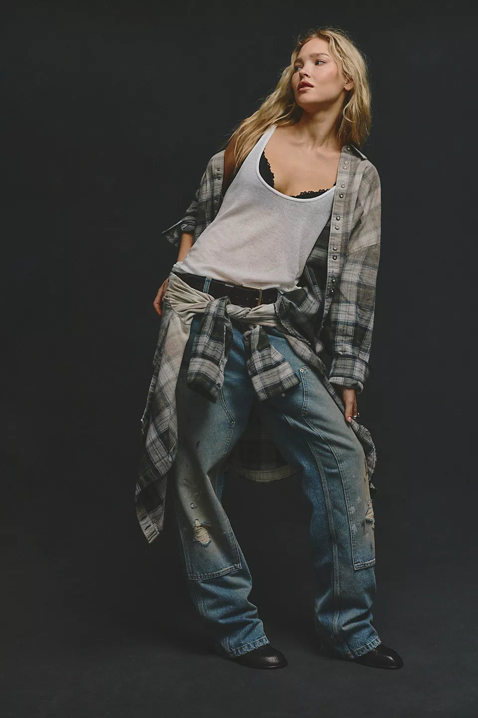 woman wearing green and beige plaid shirt open at the front with white tank top underneath. Has the same shirt tied around her waist with blue jeans and black belt. 