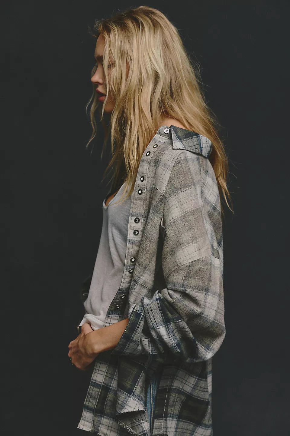 Woman modeling side view of plaid long sleeve shirt open at the front with white tank top underneath against black background. 