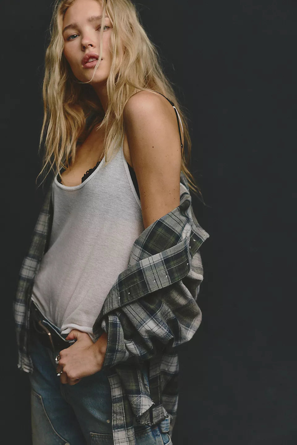 Woman wearing plaid long sleeve shirt open at the front and hanging off shoulder with white tank top underneath and blue jeans with a black belt against a black background