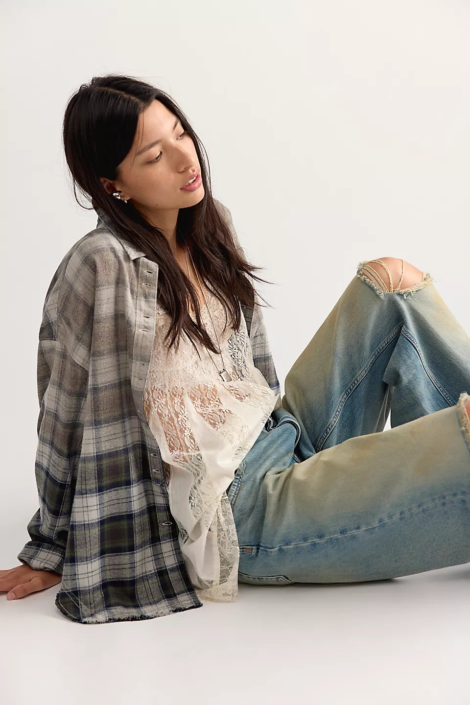 Woman sitting on floor wearing plaid, long sleeve, button down shirt with white lace shirt underneath and blue jeans.