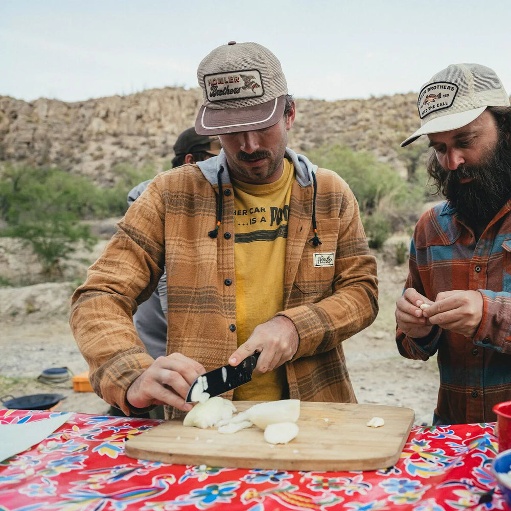Two men outdoors, one chopping onions on a cutting board with a colorful tablecloth.