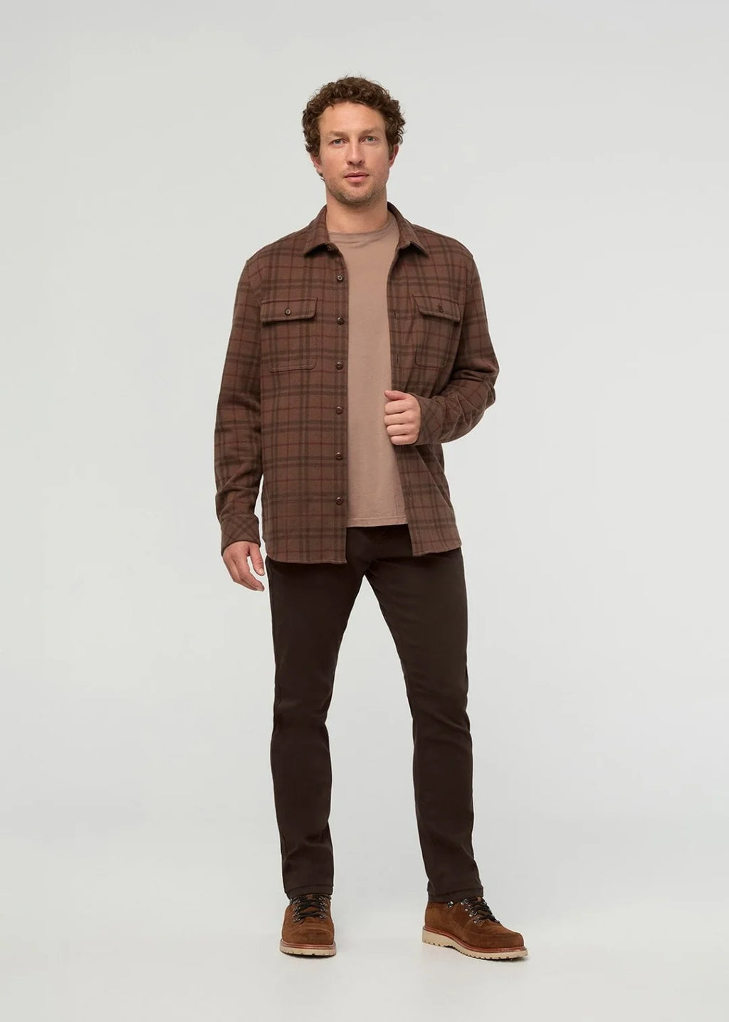 A man modeling a brown plaid button-up flannel shirt over a tan t-shirt, paired with dark brown pants and brown suede boots, against a light gray studio background.