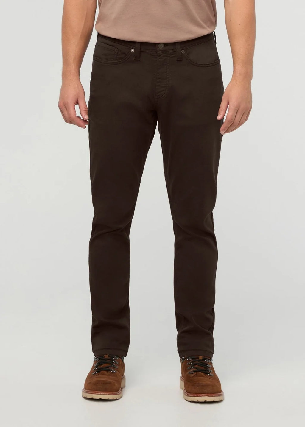 A person wearing slim-fit dark brown jeans, paired with a tan t-shirt and brown lace-up shoes, standing against a light gray studio background.