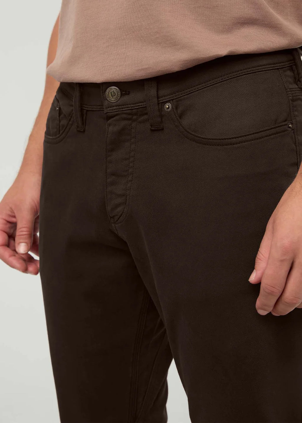 Close-up of a person wearing dark brown slim-fit jeans with a beige t-shirt, showing the front pocket and waist details.