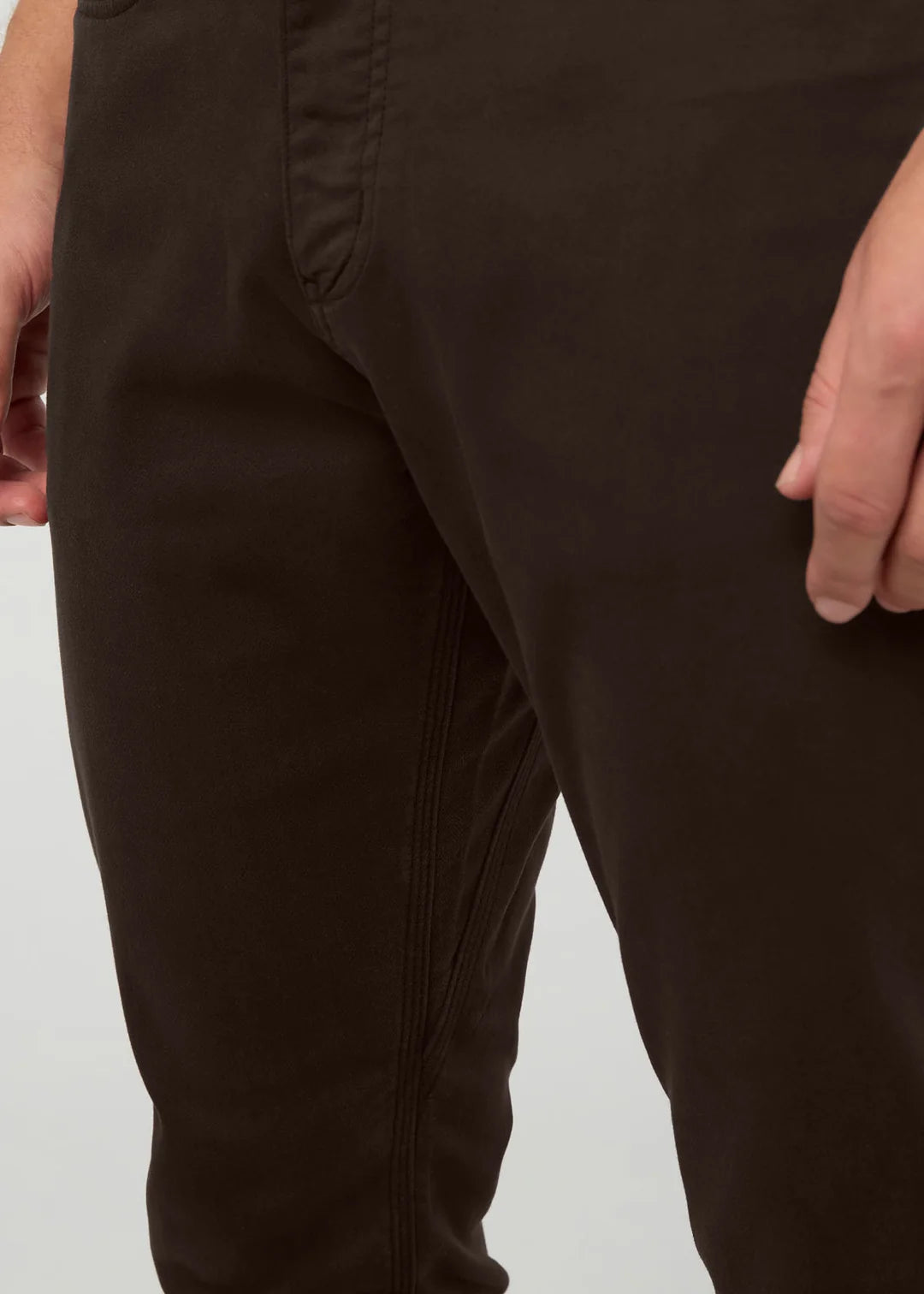 Close-up of a person wearing dark brown pants, showing the thigh area, pocket seam, and hands against a light gray background.