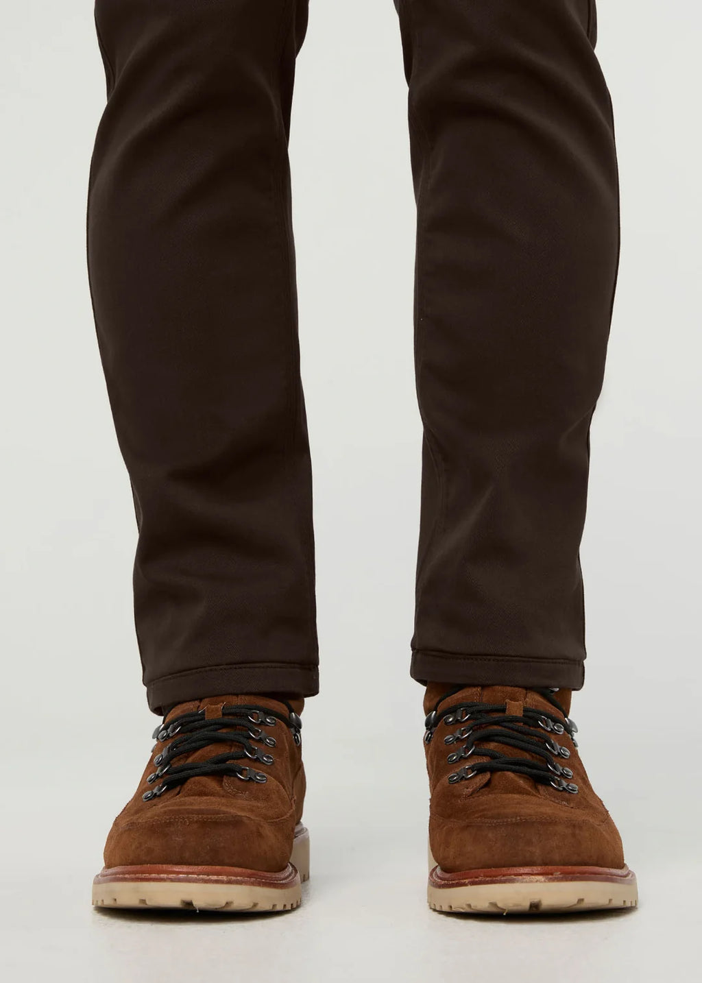 A close-up shot of a person's legs from the knees down, wearing dark brown slim-fit trousers and brown suede lace-up boots with black laces, set against a plain light gray background.
