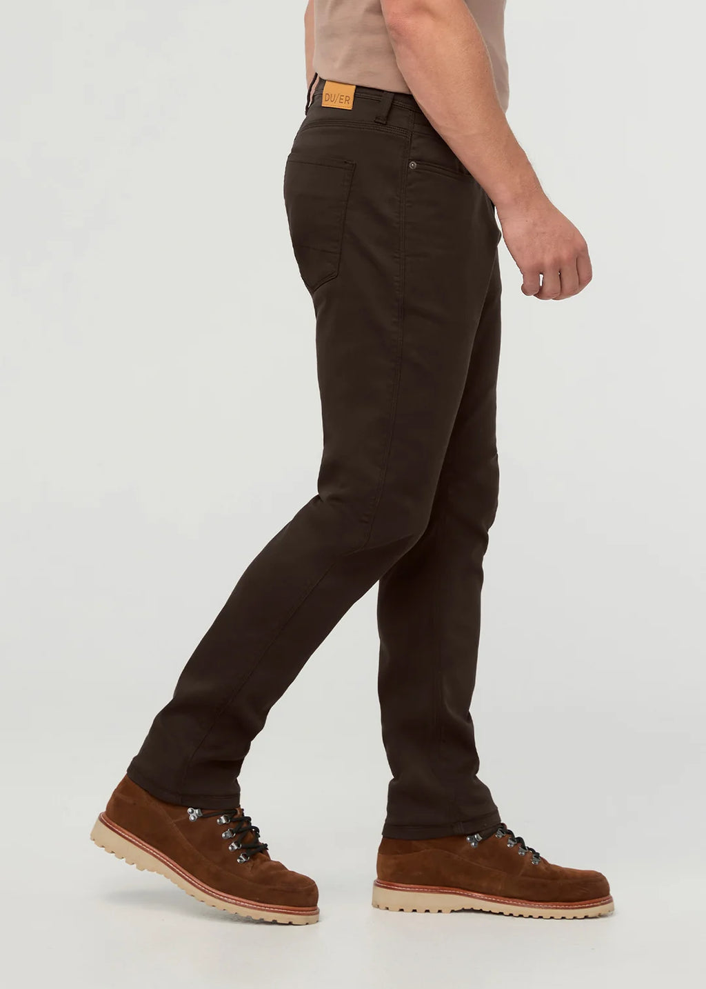 A side profile of a person wearing dark brown slim-fit jeans and brown suede lace-up boots against a light gray studio background.