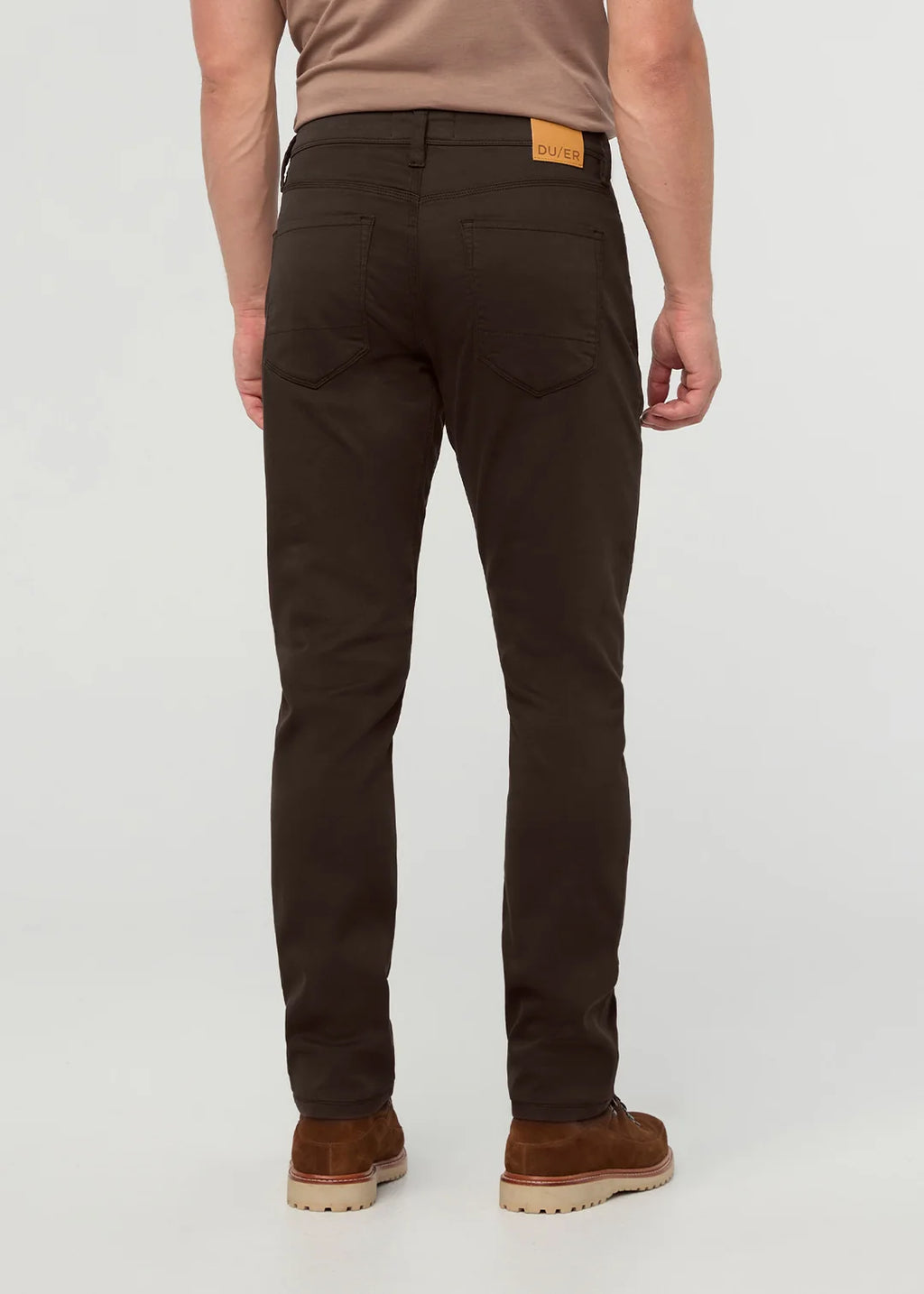 Rear view of a man wearing slim-fit dark brown trousers, a tan t-shirt, and brown suede boots against a light gray studio background.