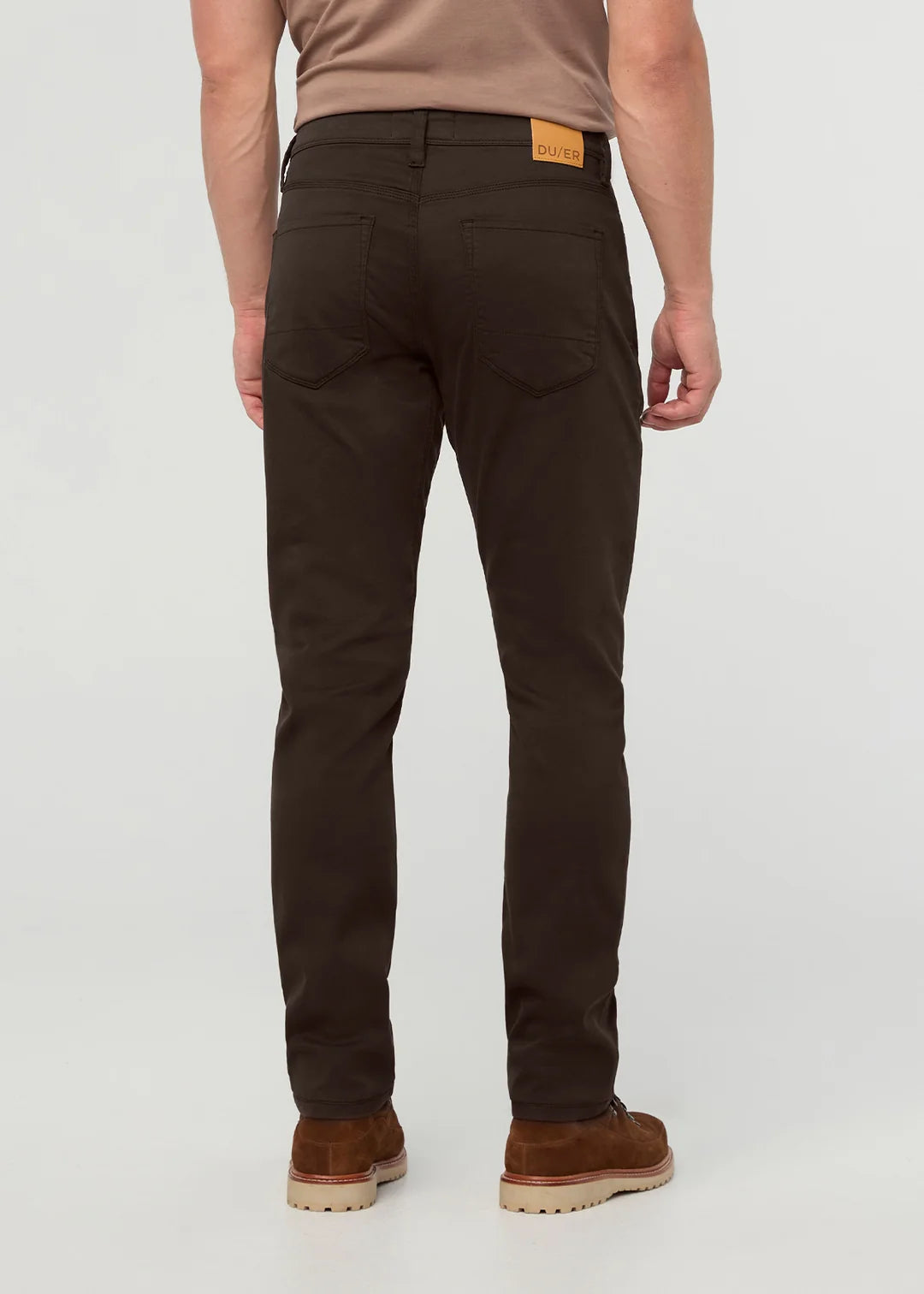 Rear view of a man wearing slim-fit dark brown trousers, a tan t-shirt, and brown suede boots against a light gray studio background.
