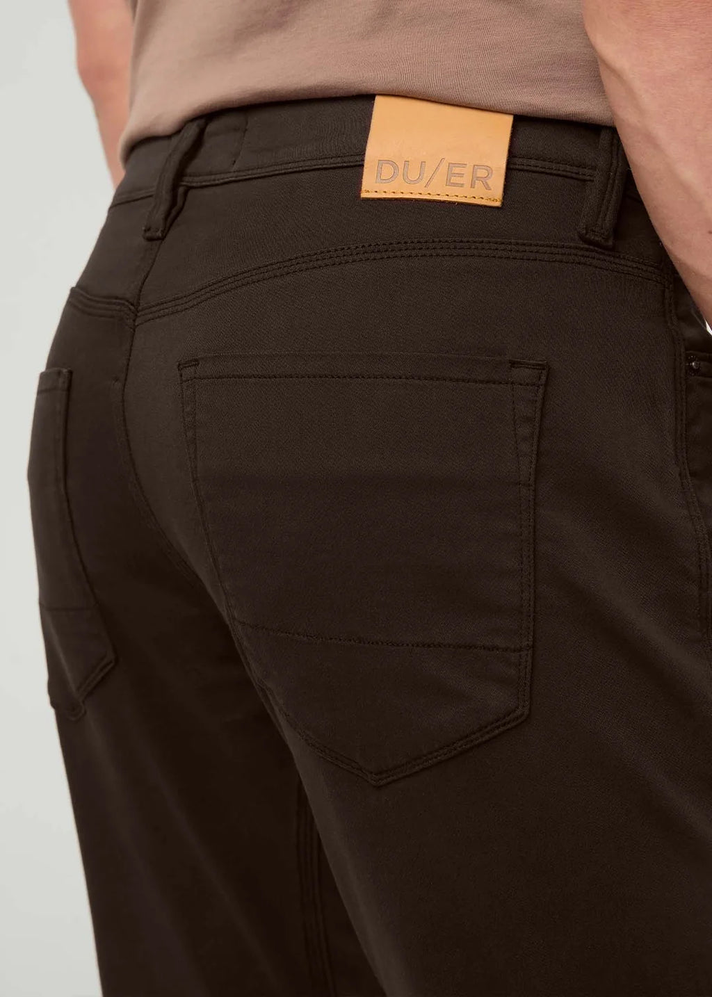 Close-up of a person wearing dark brown pants, showing the back pocket, waistband, and a tan leather DUVER brand patch.