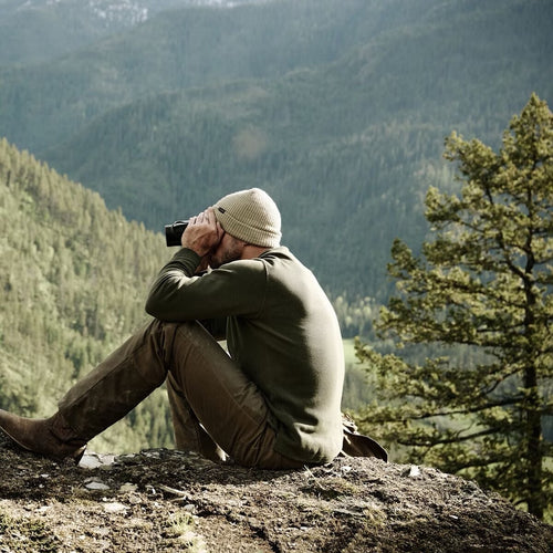 A man in an olive green jacket, brown pants, and a beige beanie sits on a rocky outcrop in a mountain landscape, holding binoculars to his eye. Dense green forests cover the mountains in the background.