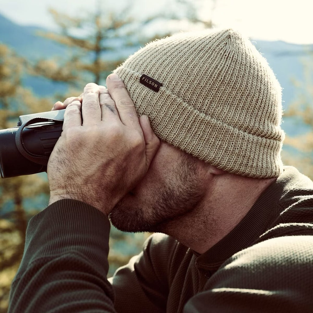 A man outdoors wearing a beige ribbed knit beanie with a small black brand patch and looking through binoculars.