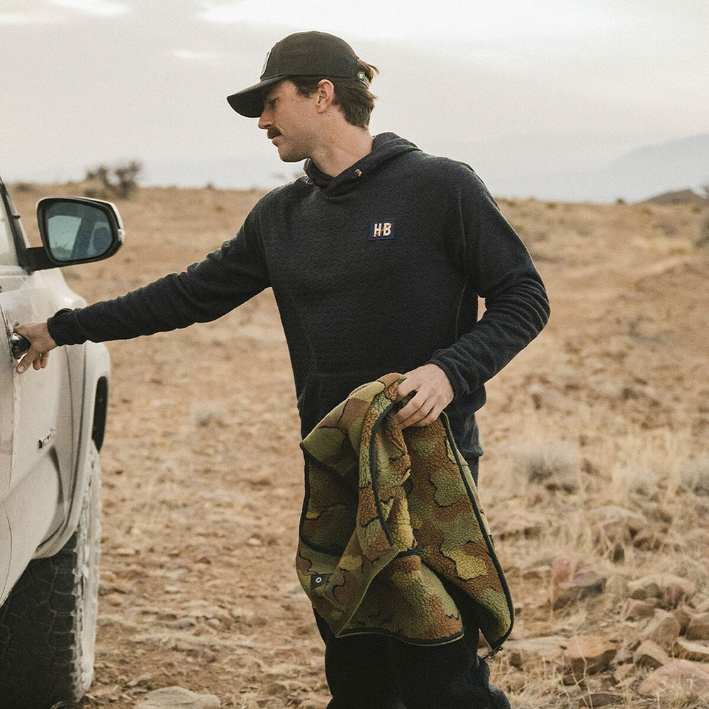 Man in black hoodie with 'HB' logo holding a camouflage blanket next to a vehicle in a desert setting